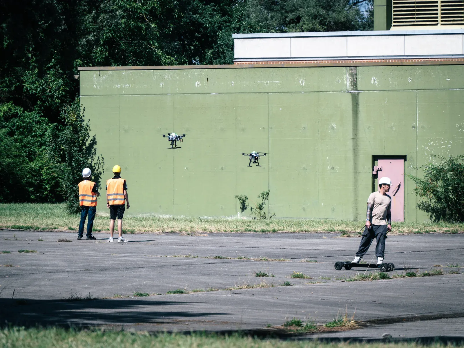 Two HERMES quadcopters hover in front of a green concrete wall while a person on an electric skateboard passes below and two team members in orange vests watch.